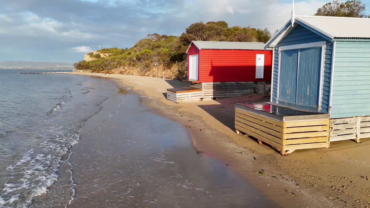 Gentle waves approach vibrant beach huts on sandy shore, soft natural light, smooth camera pan