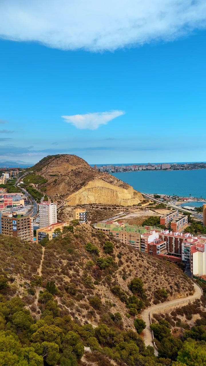 Alicante, Spanien, 8. august 2025: Forhøjet udsigt over klippefyldt bjergside og kystlinje, Alicante, Spanien. Tørt terræn med bybygninger, blåt hav og spredte skyer set ovenfra