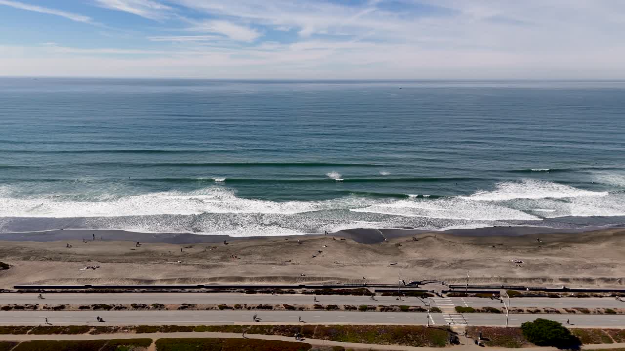 Straight-on drone shot of peaceful waves hitting the dark sandy Ocean Beach in San Francisco under soft clouds and a clear blue sky.