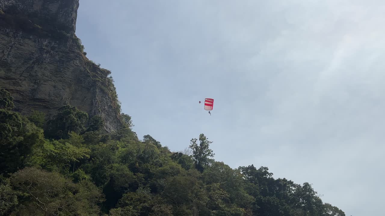 Paracadutista che scende con la calotta aperta accanto a un'alta scogliera calcarea e a una collina boscosa vicino alla spiaggia di Ao Nang, scena d'azione aerea sotto un cielo sereno a Krabi, Thailandia