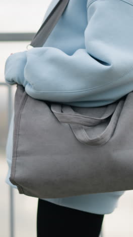 close-up view of young woman holding a handbag while walking along an iron fence, background features tranquil river view, focus on her confident stroll in casual attire