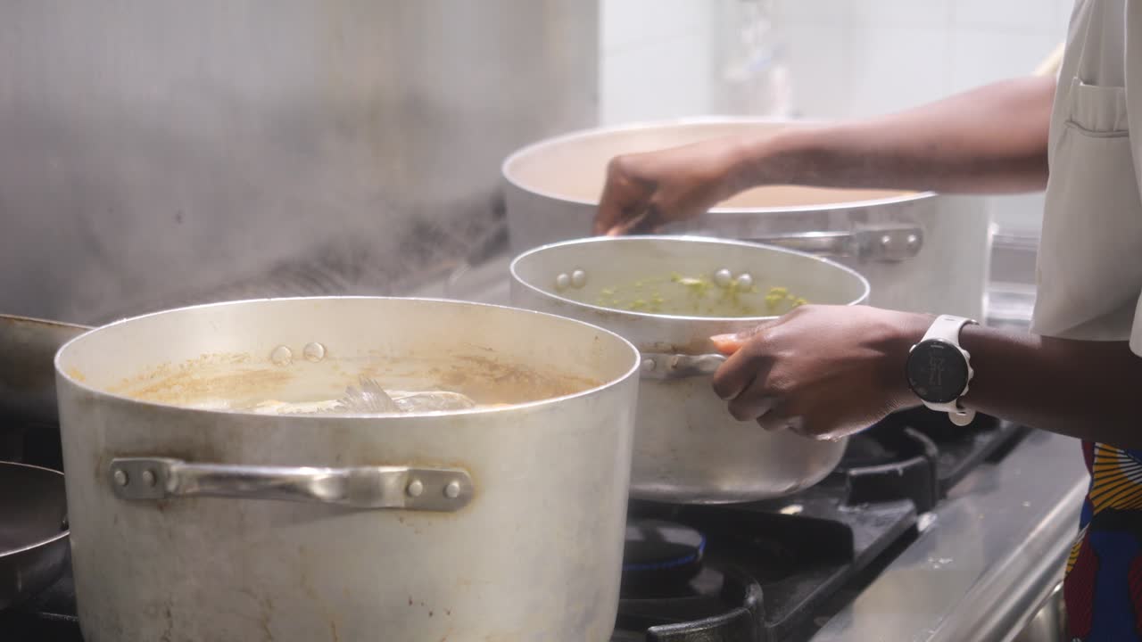 Hands of female African chef as she manages a heavy-duty commercial range, Wear white smartwatch and professional attire, reaches toward stainless steel pot to check cooking of green meal, static shot