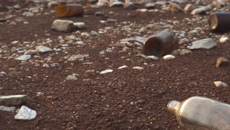 Toma-Panorámica-De-Muchas-Botellas-De-Cerveza-Marrón-Vacías-Que-Se-Tiran-En-La-Orilla-Llena-De-Rocas-Y-Guijarros-Con-Vistas-Al-Lago-Al-Fondo-En-Un-Parque-Nacional