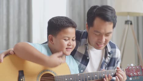 close up of asian father and son playing the guitar and singing together on sofa at home