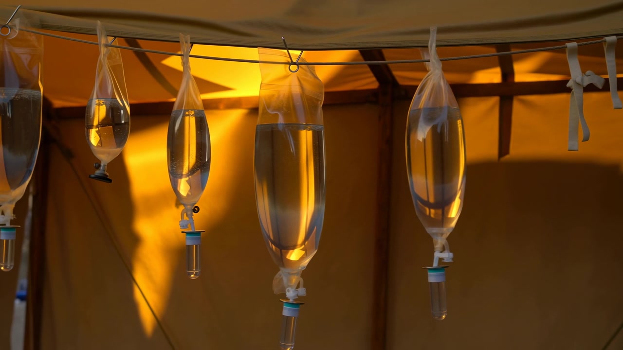 Diversi contenitori trasparenti appesi, pieni di liquido, rimangono allineati lungo un cavo mentre una luce calda attraversa le superfici traslucide, rivelando riflessi, ombre e un'ambientazione interna tranquilla