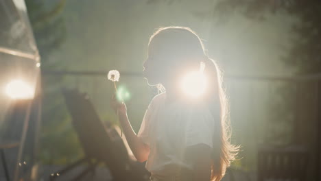 child blows seeds from dry dandelion. girl enjoys flowers standing against bright sun in yard of country house side view slow motion. vacation in natural environment