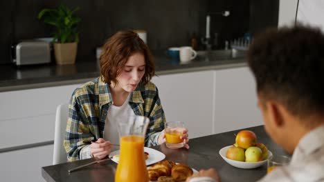 Over-the-shoulder-a-happy-young-adult-girl-with-brown-hair-and-a-bob-hairstyle-in-green-checkered-is-having-breakfast-with-her-boyfriend-with-Black-brunette-skin-color-during-a-shared-breakfast-in-the-morning-in-the-kitchen