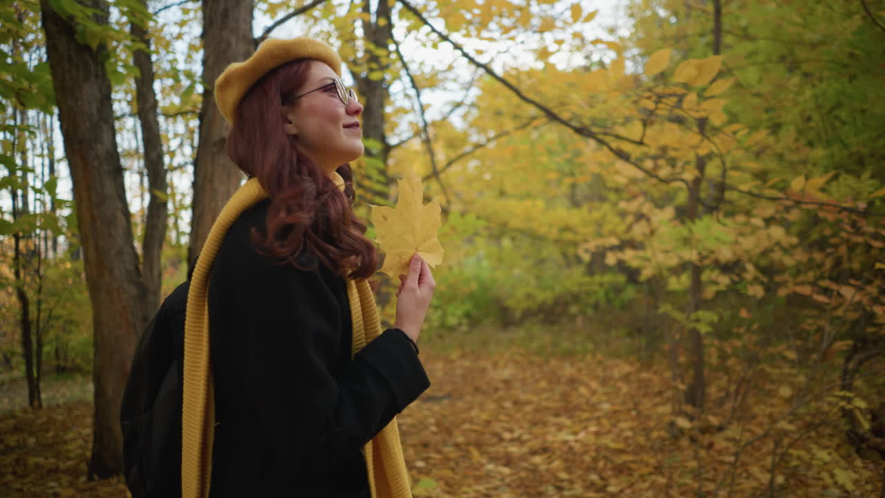 vista lateral de una mujer que lleva una bolsa con una cálida sonrisa sosteniendo una hoja de otoño jugosamente mientras camina por el bosque rodeada de follaje dorado, disfrutando de la naturaleza pacífica y el aire fresco del otoño