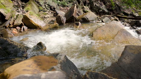 Sunny-spingtime-shot-of-water-flowing-down-a-rapid-stream-with-moss-filled-rocks