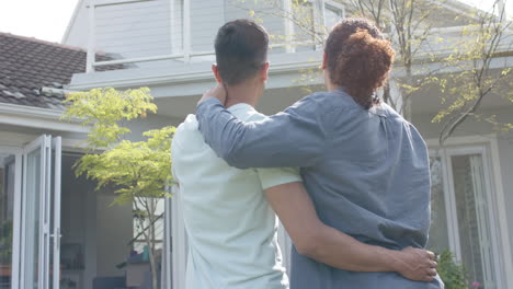 rear view of happy diverse gay male couple embracing in sunny garden of new home, slow motion