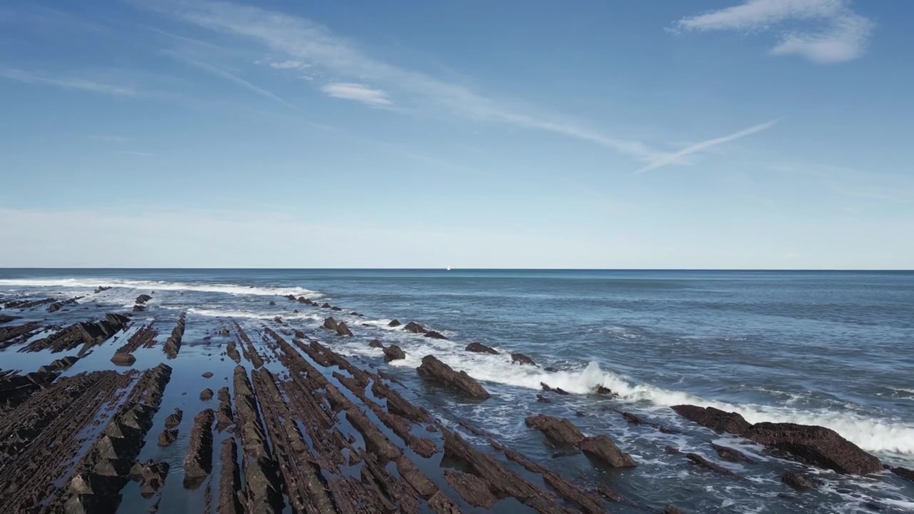 Luftfoto flyvende over de unikke Flysch-klippeformationer ved Sakoneta-stranden i Geopark Baskerlandet, mens tidevandet stiger og dækker de dramatiske geologiske mønstre med havbølger.