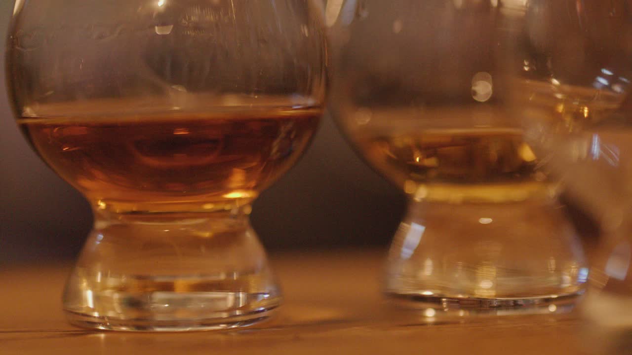A close-up sequence shows a hand arranging whiskey glasses on a wooden bar under warm, ambient lighting, emphasizing the rich amber liquid and glass reflections