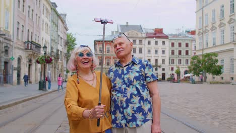 Senior-old-tourists-man-with-woman-walking-in-city-with-smartphone-on-selfie-stick-and-taking-photos