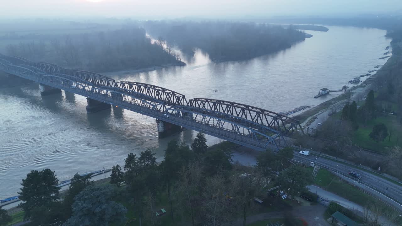 L'ampia prospettiva rivela l'orizzonte nebbioso e a basso contrasto della Pianura Padana, dove le sponde del fiume sono fiancheggiate da alberi ripariali dormienti e dalla Lombardia industriale, Italia, SS10 Strada Padana Inferiore