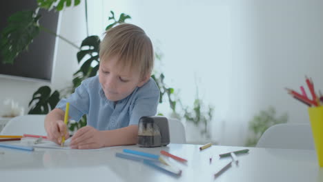 Lächelnder-Junge-Im-Blauen-Hemd-Zeichnet-Mit-Einem-Bleistift-Auf-Papier,-Während-Er-Am-Tisch-Im-Wohnzimmer-Sitzt