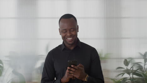 Porträt-Eines-Jungen-Afroamerikanischen-Unternehmers,-Der-Sein-Smartphone-Im-Büro-Nutzt-Und-Spaß-Daran-Hat,-SMS-Zu-Schreiben,-Auf-Dem-Mobiltelefon-Zu-Surfen-Und-SMS-E-Mails-Zu-Senden,-Die-Lächelnd-Und-Zufrieden-In-Zeitlupe-Lächeln