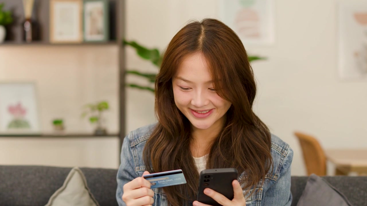 一位快乐的年轻女子坐在室内，将信用卡信息输入到她的智能手机中。