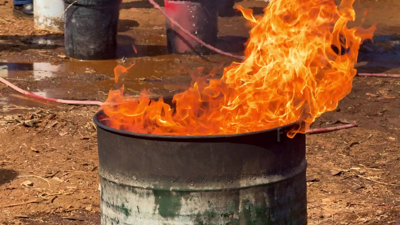 Klare flammer brænder inde i en metaltromle under brandsikkerhedstræning. Udendørs optagelser af brandfare, nødøvelse og brandbekæmpelse.