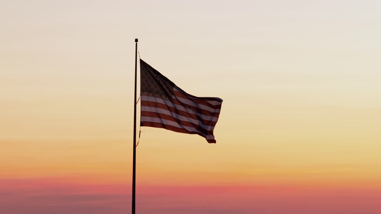 Bandera estadounidense ondeando al viento al atardecer contra un cielo colorido, capturada en vista aérea, simbolizando patriotismo, libertad y orgullo nacional en una serena atmósfera vespertina en todo EE. UU.