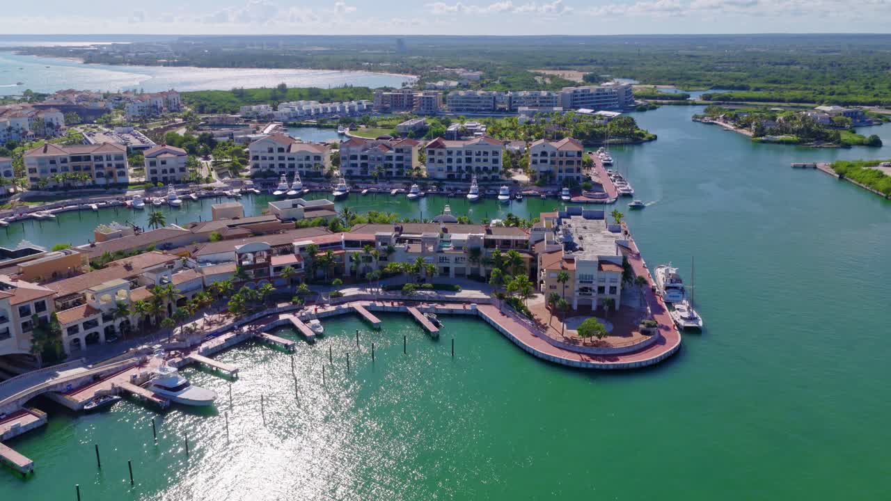 Luftaufnahme von Cap Cana Marina in der Dominikanischen Republik mit türkisfarbenem Wasser, Luxusjachten, Wohnhäusern am Wasser und von Palmen gesäumten Gehwegen. Lebhafte tropische Küstenszene. Weitwinkelaufnahme