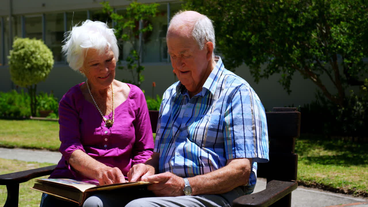 vista frontal de una pareja de ancianos caucásicos activos mirando un álbum de fotos en el jardín del hogar de ancianos 4k