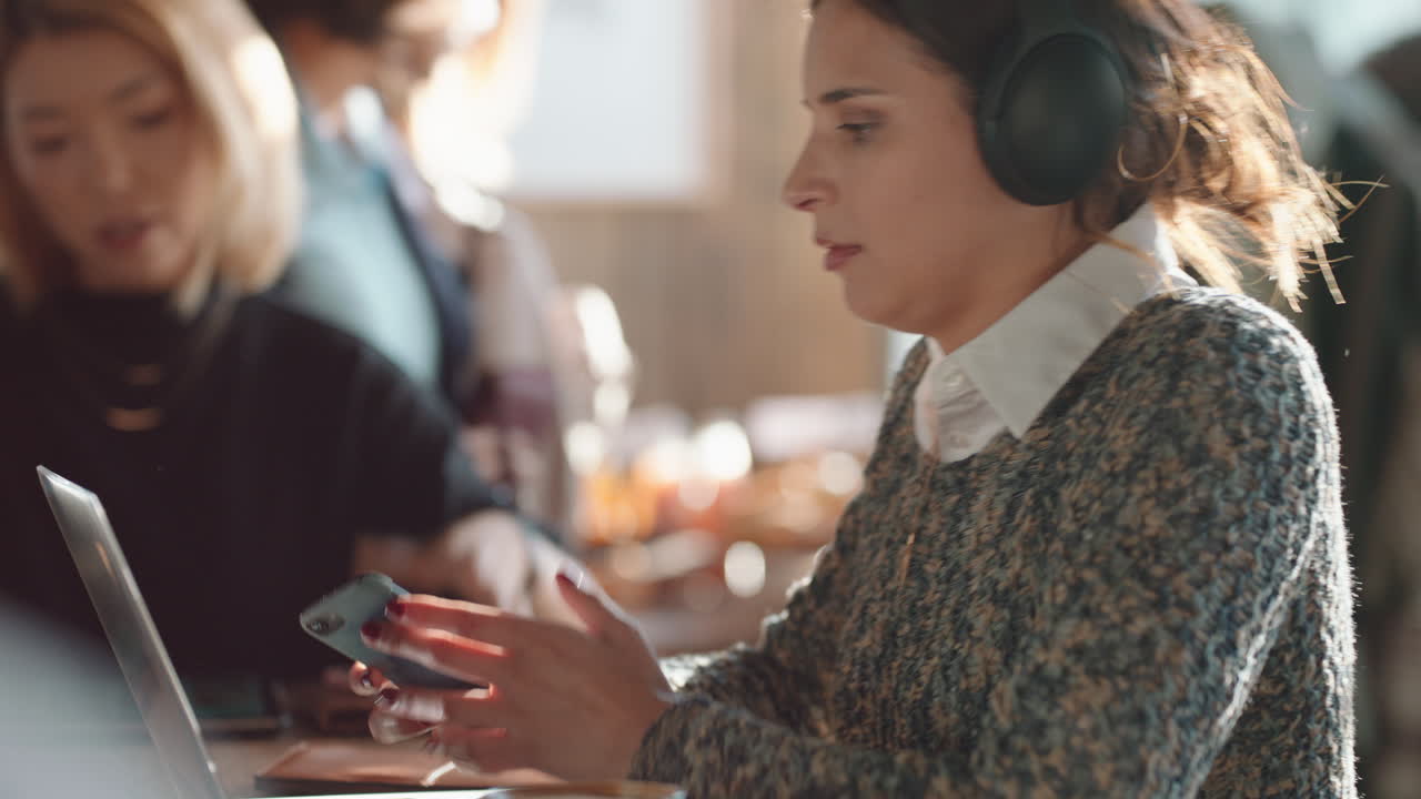 young woman using laptop working in cafe typing email sharing messages on social media wearing headphones enjoying listening to music in busy restaurant drinking coffee