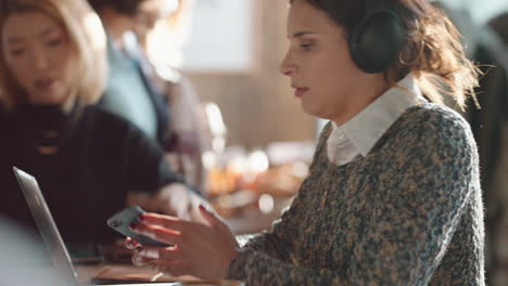 young woman using laptop working in cafe typing email sharing messages on social media wearing headphones enjoying listening to music in busy restaurant drinking coffee