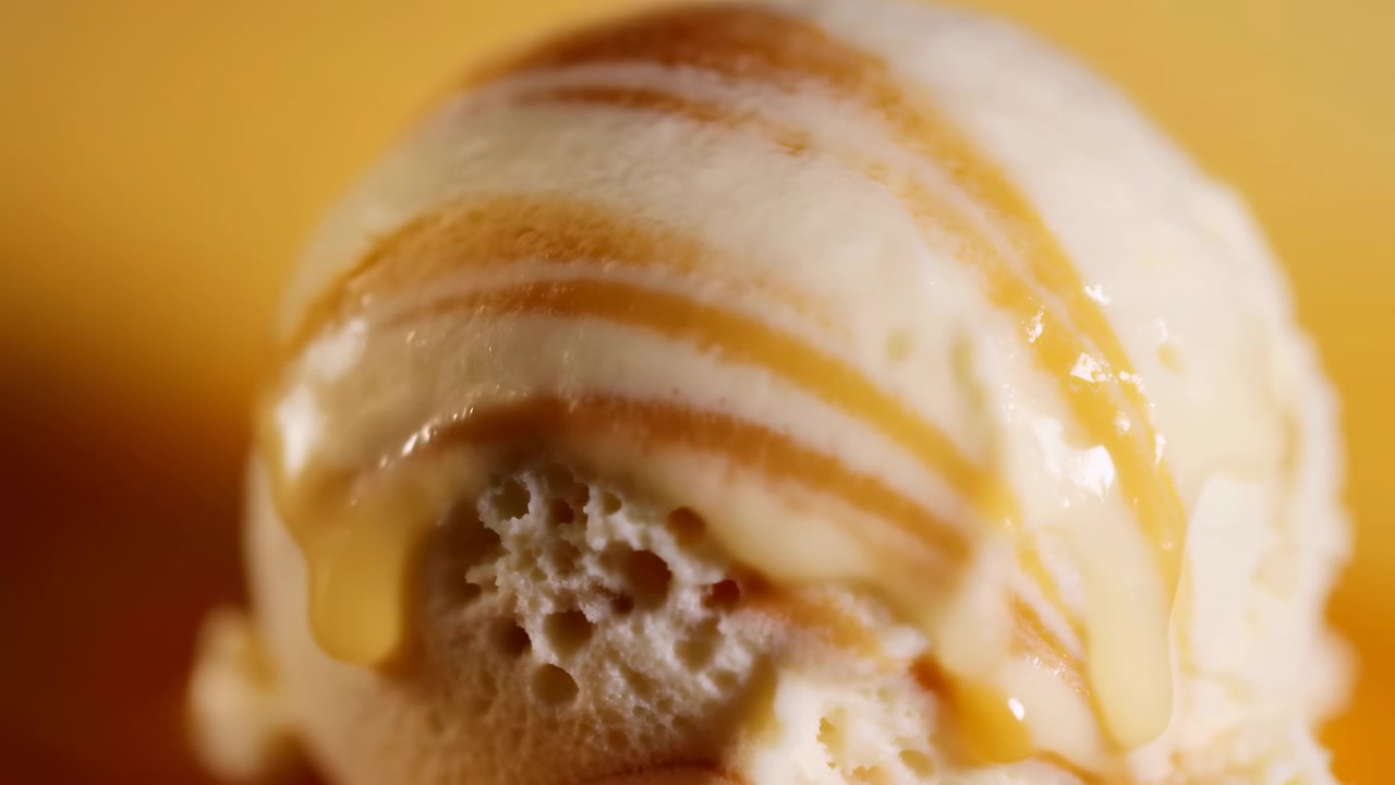 Un movimento macro mostra salsa al caramello versata su una pallina di gelato alla vaniglia, creando nastri lucidi, sottili scie, piccole goccioline e un leggero accumulo mentre lo sciroppo scorre su morbide creste con riflessi caldi