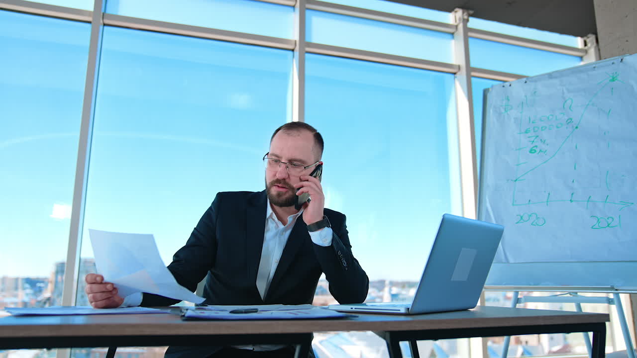 Serious businessman working in office. Bearded man boss in suit sitting at the desk with a laptop and talking the phone. Business concept.