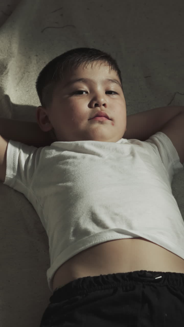 운동 후 체육관에서 머리 에 손을 고 누워있는 어린 아시안 소년은 무술 클럽에서 느린 동작으로 휴식을 취합니다. 주니어 스포츠맨은 바닥 개에 편안하게 있습니다.