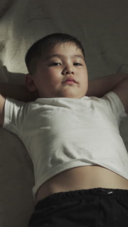 niño se acuesta con las manos debajo de la cabeza en el gimnasio después del entrenamiento. pequeño niño asiático disfruta del descanso en el club de artes marciales en cámara lenta. deportista junior se relaja en la cubierta del suelo
