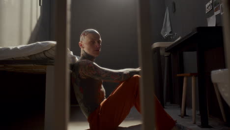 portrait of bald tattooed inmate sitting on floor in prison cell