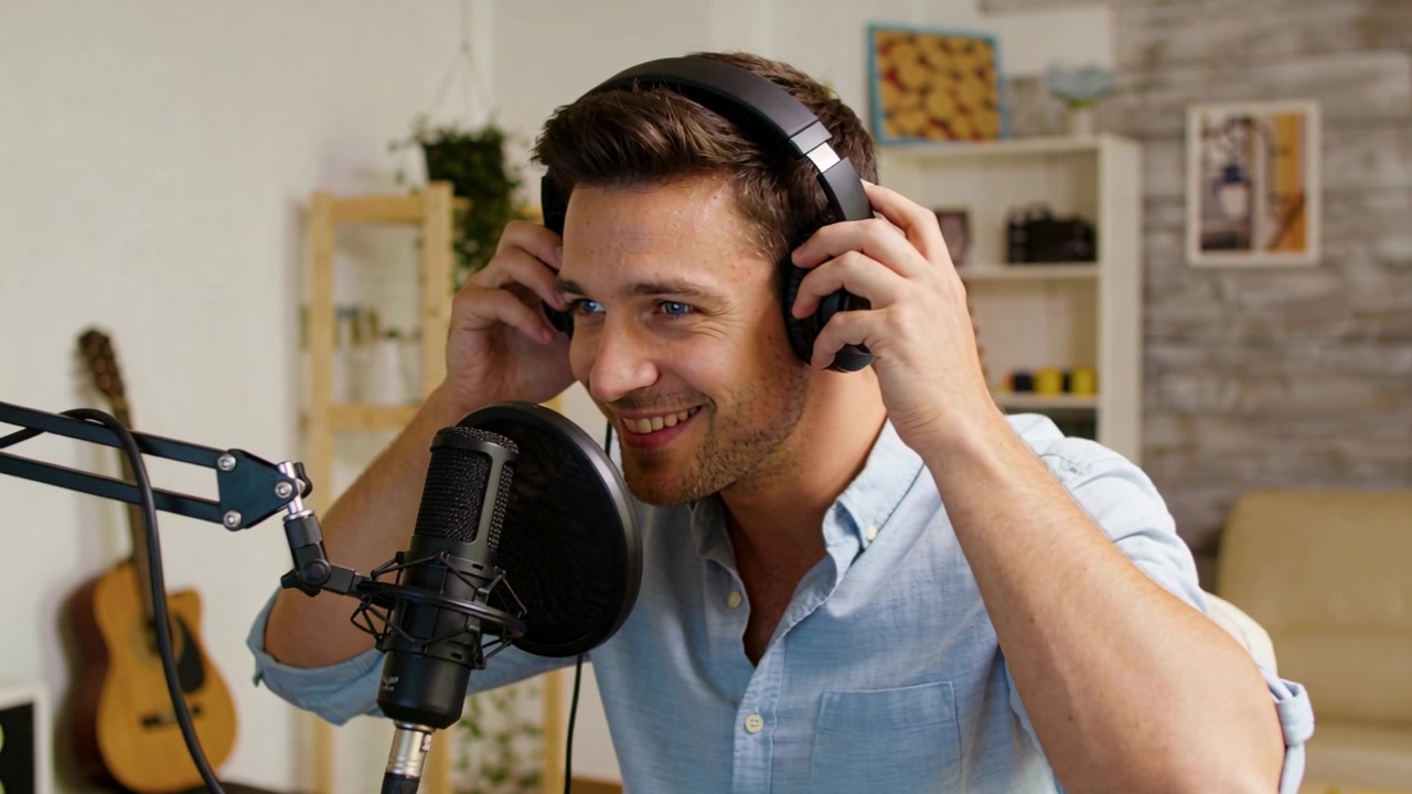Uomo sorridente che solleva le cuffie, regolandole e chinandosi verso il microfono in studio, preparandosi a registrare.