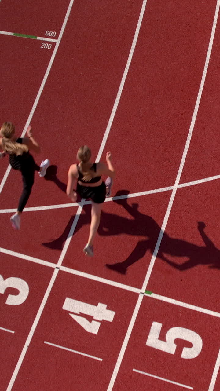 Perspectiva a vista de pájaro de dos mujeres trotando por una pista de atletismo sintética roja en un estadio deportivo. Ideal para contenido de fitness, competición y estilo de vida saludable.