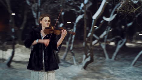 a gorgeous girl in a white dress with sparkles and in a fur coat is standing in the park and playing the violin in the winter on the background of trees in the evening. camera motion to right