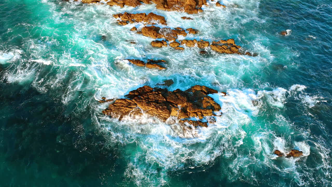 Some rocks stick out of blue water of the sea or ocean. Waves splash at the rocks. Aerial perspective.