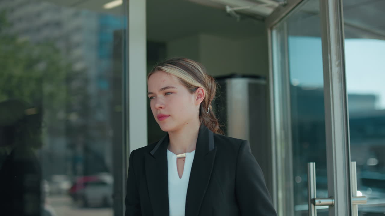 Professional designer opens glass door walking out of building with her reflection visible on door surface, parked cars and trees lining urban street in background