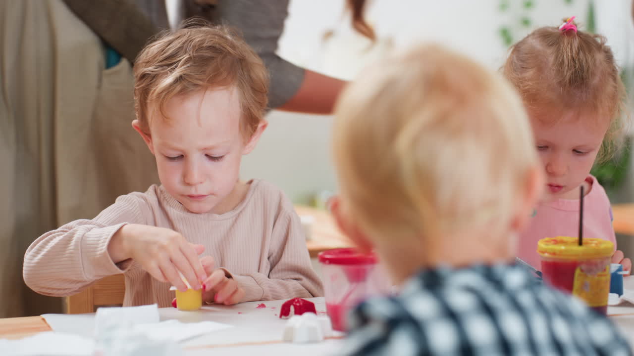 Little boy in beige top focuses on opening paint container during art class, red paint staining his thumb while other children engage in creative activity around table under teacher guidance indoors