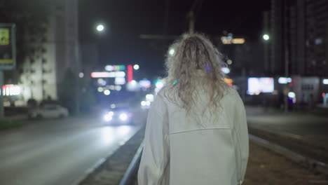 rear view of tired woman in white coat walking slowly along rail track at night while car with headlights passes on city street surrounded by glowing signs and blurred urban buildings