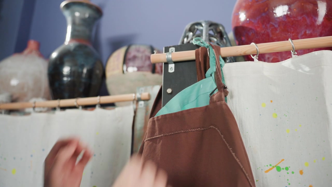 Seniman merapikan celemek sebelum pelajaran. Seniman keramik menyesuaikan celemek saat sinar matahari menerangi area kerja. Studio tembikar yang semarak dengan seniman mengikat celemek di antara vas-vas berlapis glasir dan cahaya matahari yang hangat.