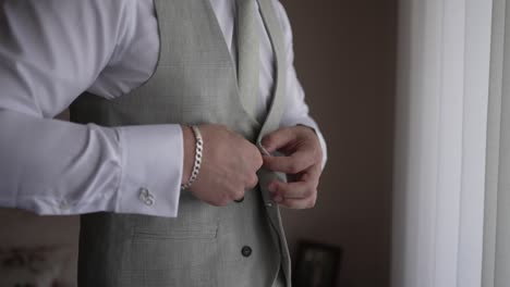 Man-buttoning-his-grey-vest-while-wearing-a-white-dress-shirt-and-tie,-near-a-window