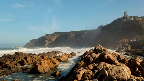 Wellen-Schlagen-In-Zerklüftete-Riffe-Am-Poort-In-Mossel-Bay,-Blick-Auf-Den-Leuchtturm