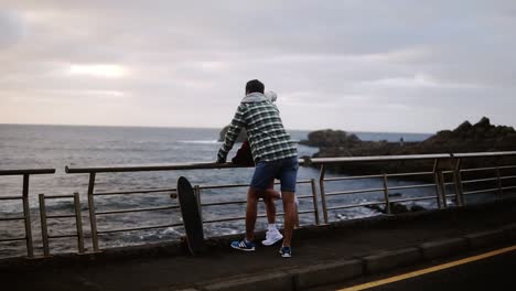 Woman-and-standing-together,-looking-on-ocean---Young-couple-shown-from-the-side-look-out-over-an-ocean-in-front-of-the-hills,-talking,-embracing.-Cloudy-weather.-Loving-couple