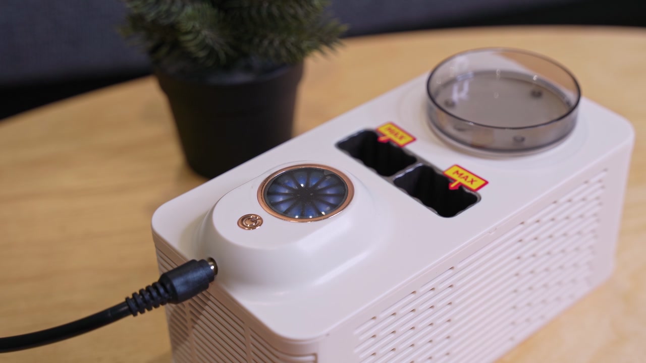 Close-up of a compact white electronic appliance with a power cable, vented housing, circular controls, labeled slots, and a clear round compartment on a wooden table indoors.