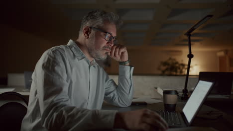 exhausted senior man rubbing eyes as working on laptop late in office