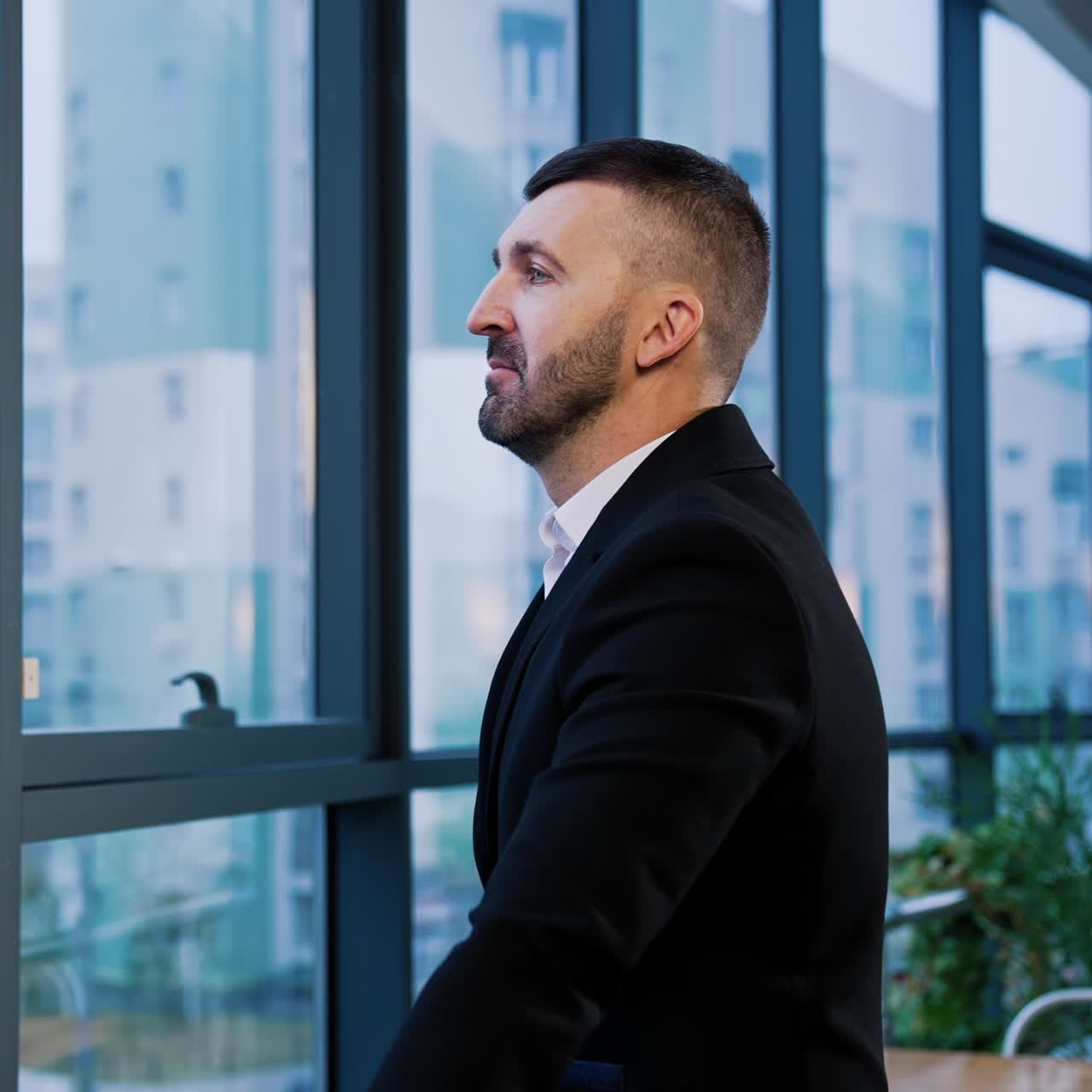 Mid-aged bearded businessman in black suit in the office. Serious confident man stands looking at the window
