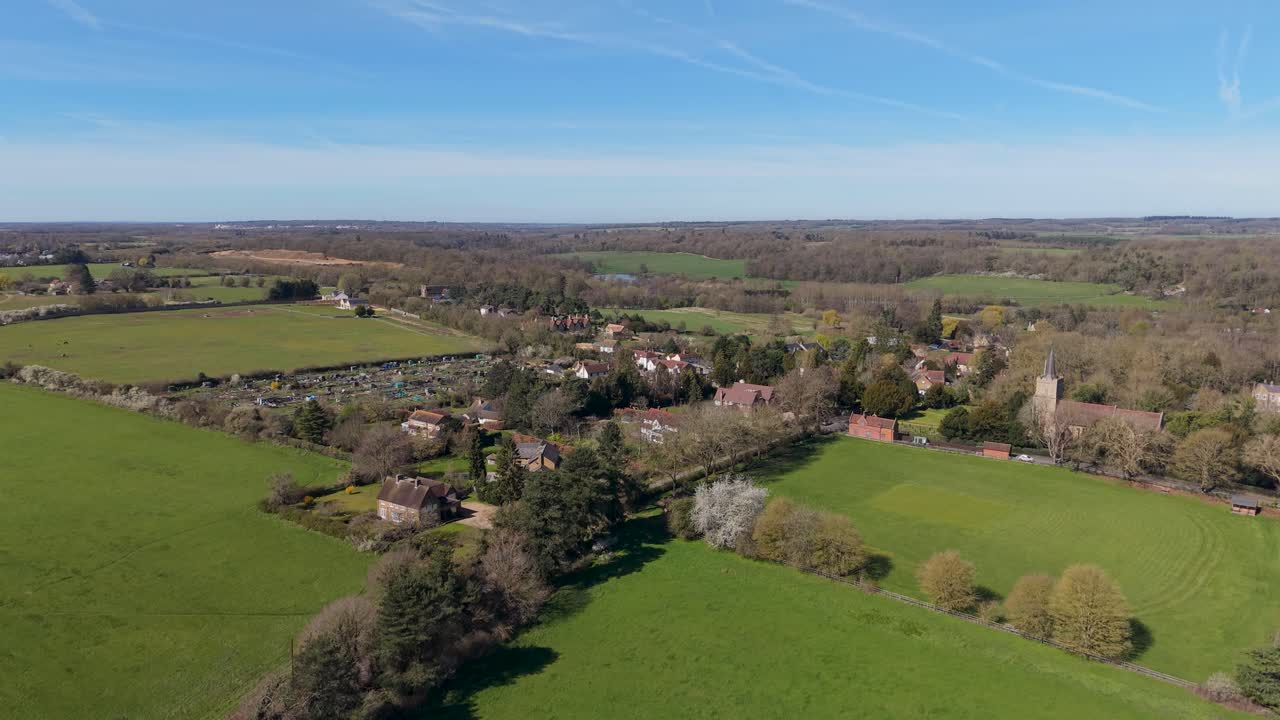 Hertingfordbury village aerial descent reveals the full rural settlement, church, fields and woodland under a clear blue sky, showcasing Hertfordshire countryside in England