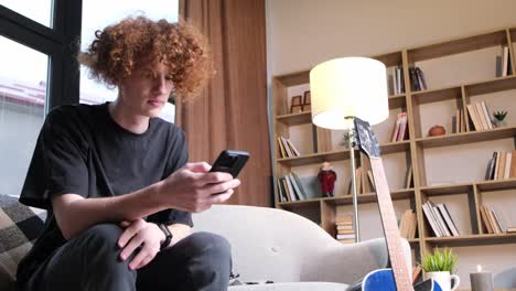 a guy with curly red hair is looking for something on the phone on the internet a guy with curly red hair is looking for something on the phone on the internet in a modern living room young modern guy and gadgets
