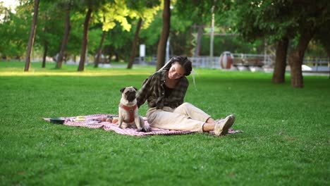 smiling girl sitting on plaid on lawn in a park., the dog sitting in next to her, she is wondering, looking at him