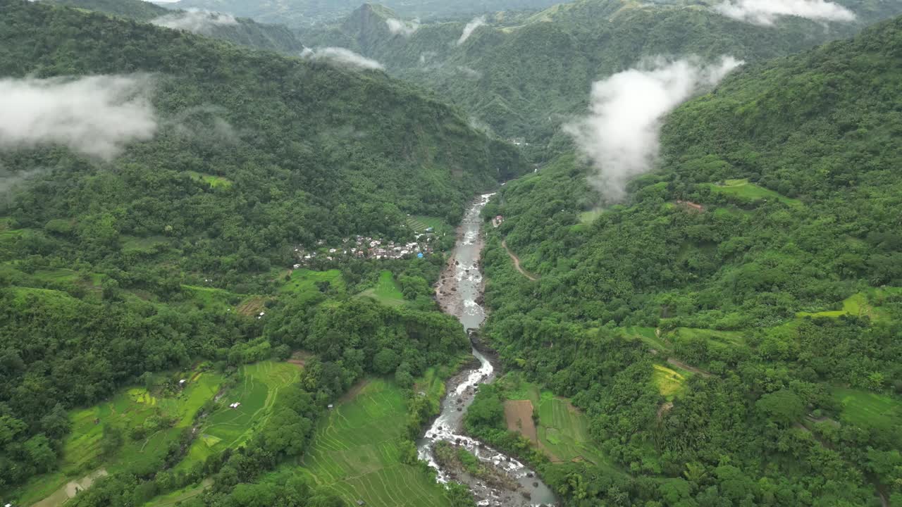 A wide aerial angle drifts to a final hold over forested hills and a winding river. Mist threads through valleys as cultivated land and a small village settle into the lush green terrain